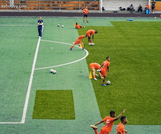 A group of people wearing bright orange sportswear are practicing on a green field. They appear to be athletes, likely soccer players, stretching and preparing for training. There are several soccer balls on the field, and a coach is observing from a distance. The field is surrounded by bleachers with a few onlookers seated, and the area is bordered by trees and residential buildings in the background.