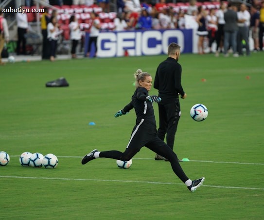 A soccer player in a black sports outfit is mid-action on a green field, focusing intently on kicking a soccer ball. Several soccer balls are scattered on the grass nearby, and a coach or teammate stands in the background watching. Spectators can be seen in the distant stands.