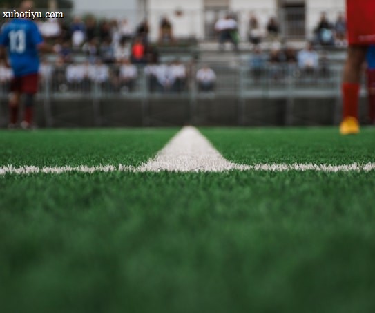 A close-up view of a green soccer field's artificial turf, focusing on a white dividing line running down the center. In the blurred background, there are players wearing sports uniforms and spectators seated in stands.