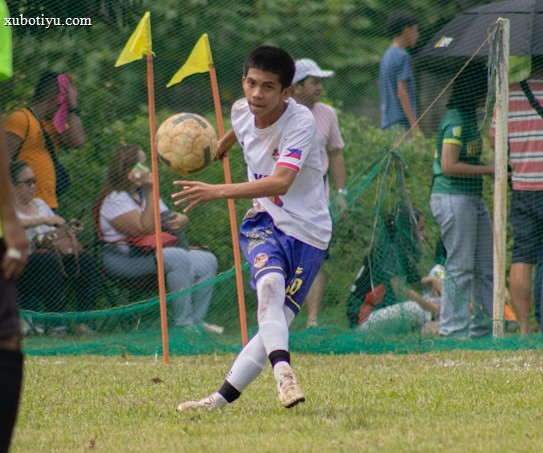 A soccer player in a white and blue uniform is seen in action on the field, focusing intently on controlling the ball with precision. Behind him, spectators watch from the sidelines, seated or standing near a net barrier. The setting indicates an outdoor event on a grassy pitch with vibrant green surroundings.